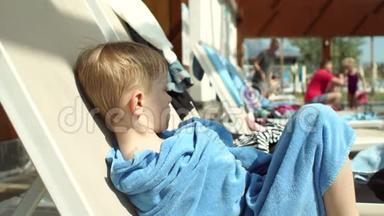 一个悲伤的小男孩在游泳池旁的休息室上的肖像，他被裹在毛巾里。