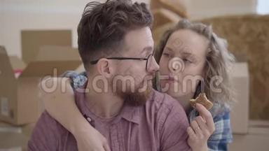 漂亮女人吃饼干的肖像，从背后拥抱戴眼镜的胡子男人。 未包装的箱子在