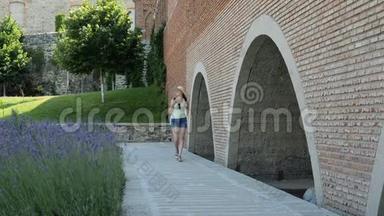戴着一顶背包的年轻女人沿着古老的墙壁走着，摸着她的手。 旅行和建筑