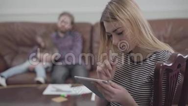 美丽的金发女人坐在丈夫和小女孩模糊的身影面前，手里拿着平板电脑的画像