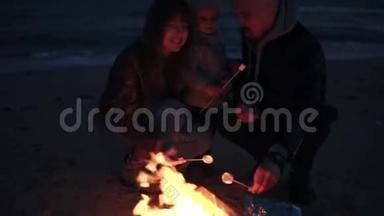妈妈，爸爸穿着暖和的衣服，正在用木棍和他们的小宝宝烤棉花糖。 海滩上的篝火