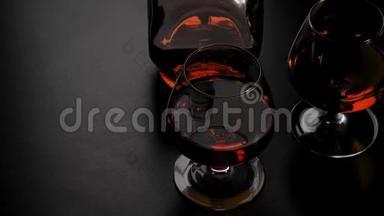 豪华白兰地。 带有金色干邑的圆形瓶子和两个玻璃杯在黑色背景下的黑色桌子上旋转。 玻璃眩光。