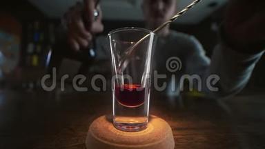 酒保将透明的酒精倒入分层的镜头中，用吧勺缓慢地用红色的酒，将酒精倒入一个