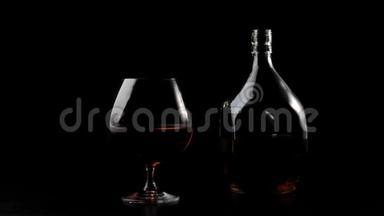 豪华白兰地。 一个方形瓶子里的金色干邑站在黑色桌子上的玻璃旁边，背景是黑色的。 玻璃眩光。