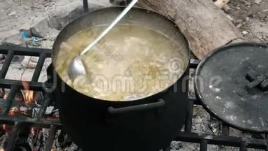 一只手拿着一个瓢，把配料混合在沸腾的汤里。 在大自然的大锅里做饭。 徒步旅行者的食物。 营地游客