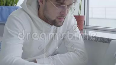 一个穿着白色运动衫、坐在桌子旁的年轻时尚、有魅力的留胡子的人，在笔记本电脑上打字
