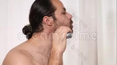 英俊的年轻人留着长发，在浴室里用<strong>修剪器</strong>刮胡子。 光线优美，背景模糊..