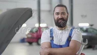 一位满脸笑容的留胡子的机械师在一家汽车修理厂的背景下摆着一把扳手的画像