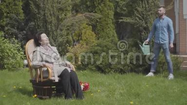 一位成年妇女坐在草地上的<strong>摇椅</strong>上享受阳光。成年孙子要来浇水了
