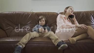 小可爱的男孩坐在皮革沙发上，两腿分开，用遥控器在电视上换频道，而姐姐则用遥控器