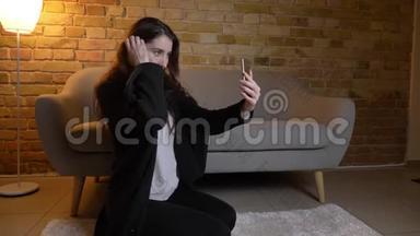 年轻的白种人女孩留着波浪发，坐在地板上，在舒适的家里，在智能手机上快乐地制作自拍照片