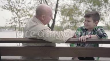 肖像祖父和孙子坐在河边的公园里的长凳上，老人讲着有趣的故事给人听
