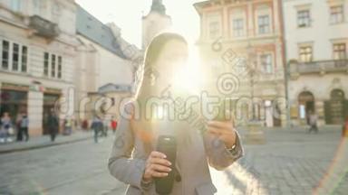 女人手里拿着一个<strong>保温</strong>杯，在夕阳下用智能手机沿着一条老街走着。 通信、社交网络