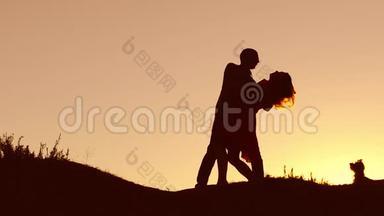 恋爱中的情侣在日落和接吻时跳舞剪影。 爱的男人和女人与狗跳舞剪影缓慢
