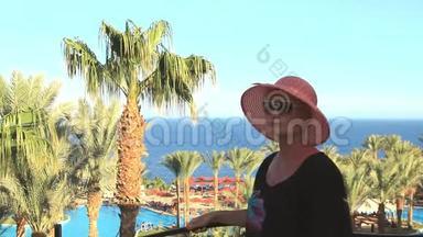 一个阳光明媚的夏日，戴着帽子的年轻美女从阳台望向<strong>蔚蓝</strong>的<strong>大海</strong>和棕榈树