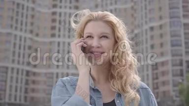 在摩天大楼前用手机说话的可爱微笑自信的金发女人的特写肖像。 城市生活方式
