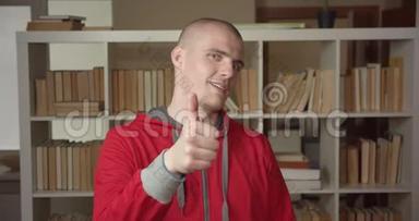 一幅年轻有魅力的白种人男学生的特写肖像，他竖起大拇指看着大学图书馆里的摄像机