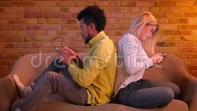 非洲男孩和白人女孩背<strong>靠背</strong>地坐在<strong>沙发</strong>上看着家里的智能手机。