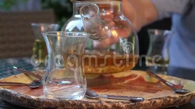 一个人把热茶从玻璃茶壶倒入一个透明的杯子里。 草药茶
