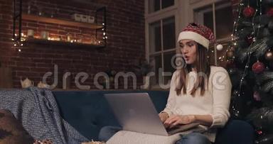 漂亮的年轻女子戴着桑塔帽，坐在<strong>家中</strong>圣诞树附近，背景是她用笔记本电脑发短信打字