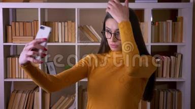 美丽的黑发女教师眼镜制作自拍智能手机在图书馆。