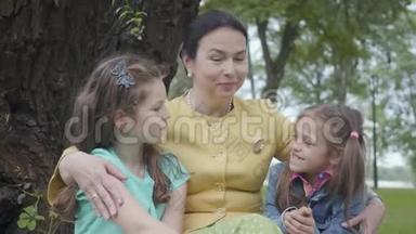肖像优雅的老奶奶坐在公园树下的草地上，拥抱着两个可爱的孙女