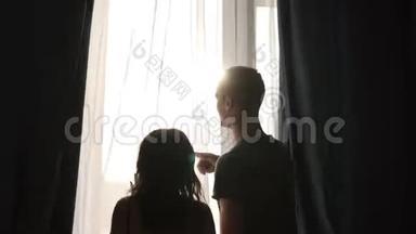 幸福的年轻夫妇穿着<strong>家居</strong>服走到窗前，拉开黑暗的<strong>窗帘</strong>。 白人男女后视拥抱