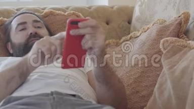 一个留着胡须的人，在柔软的米色沙发上玩智能手机游戏，在失败和被抛弃之后