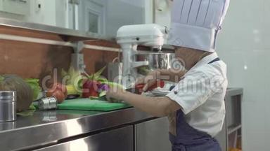 小厨师穿着围裙和厨师帽在学校厨房<strong>做饭</strong>。 有趣的小男孩<strong>做饭</strong>切新鲜蔬菜