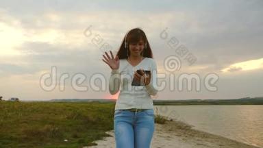 戴着耳机的漂亮女孩带着平板电脑在海滩上散步，并且正在在线视频会议。 快乐的女人在海滩散步