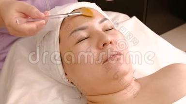 一位专业的美容师用一种滋养海藻的<strong>面膜敷</strong>在一个亚洲女人的脸上，用刷子。 复壮程序