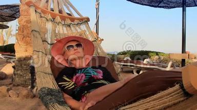 美丽的年轻女子戴着帽子，在阳光明媚的夏日棕榈树的树荫下，躺在海滨的吊床上