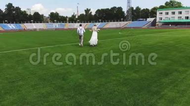 空中视频，在体育场，在一个绿色足球场的中心，新婚快乐的新婚夫妇正在跑步