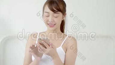 美丽的肖像年轻的亚洲女人微笑着坐着看着智能手机阅读社交网络与享受床上卧室