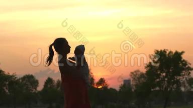 夕阳的剪影：年轻的母亲抱着她的小男孩在城市公园站在夕阳面前，生动活泼
