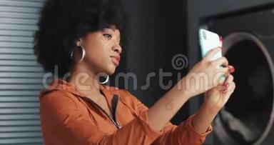快乐迷人的非裔美国年轻女子在自助洗衣店智能手机上自拍。 自助公共洗衣店。
