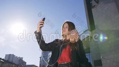年轻漂亮的金发女孩在城市街头行走时在智能手机摄像头上自拍照片的画像