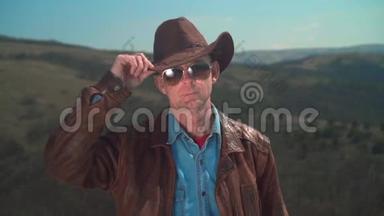 在山上，一个男人戴着牛仔帽，<strong>皮夹克</strong>，眼镜。 一个人用手摸他的帽子。