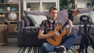 成功的年轻男子vlogger正在<strong>录制</strong>视频，弹吉他，坐在家里的地板上唱歌，并使用<strong>相机</strong>