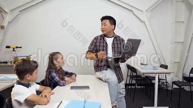 韩国男老师坐在桌子上，手里拿着笔记本电脑给学生上课。 学龄儿童认真倾听