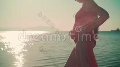 一个穿着红色一件泳衣的怀孕女孩站在海边抚摸她的肚子。 做得很好。