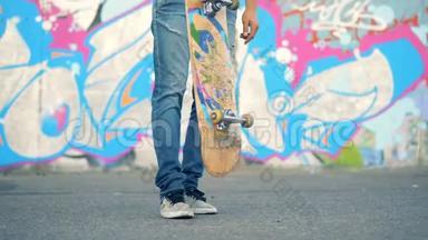 一个年轻人`站在涂鸦墙边，抬着<strong>滑板</strong>