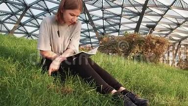 一个穿着灰色衬衫的少女坐在公园的草地上，在阳光明媚的日子里读着一本带有讲稿的笔记本