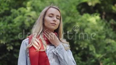 生病的年轻女子戴着红领巾，<strong>感觉不好</strong>，喉咙痛，站在公园里。 痛苦