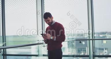 快乐的商业工作者站在一个大窗户旁边，同时在他的智能手机里输入一些东西