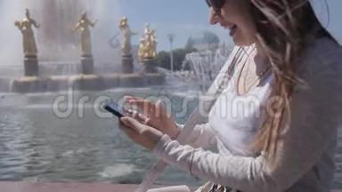 在<strong>炎热</strong>的<strong>夏天</strong>，穿着米色西装、坐在公园喷泉旁的智能手机的快乐女孩
