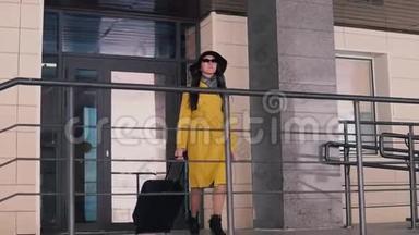 一位身穿黄色斗篷、戴着帽子、留着深色长发从酒店的门廊走出来，摘下眼镜