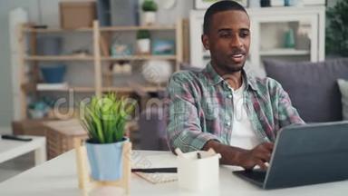非裔美国人在家工作时使用笔记本电脑打字，然后在纸上做笔记，坐在办公桌前喝酒