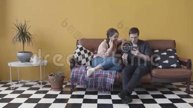 一对夫妇坐在沙发上，带着电子平板电脑，在一黄色的墙壁和<strong>黑白格子</strong>地板