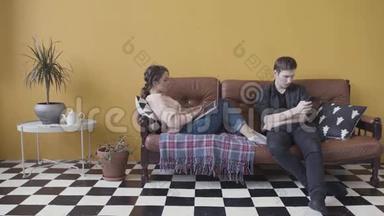 年轻夫妇坐在家里的沙发上，沉迷于现代小玩意。 库存录像。 使用手机的人